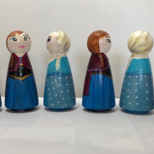 May include: Set of seven wooden peg dolls, painted to resemble characters from the film Frozen. The dolls are dressed in blue and purple gowns with white accents. Two of the dolls have blonde hair, two have red hair, and two have brown hair. There are also two small wooden snowman dolls.