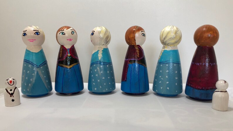 May include: Set of seven wooden peg dolls, painted to resemble characters from the film Frozen. The dolls are dressed in blue and purple gowns with white accents. Two of the dolls have blonde hair, two have red hair, and two have brown hair. There are also two small wooden snowman dolls.