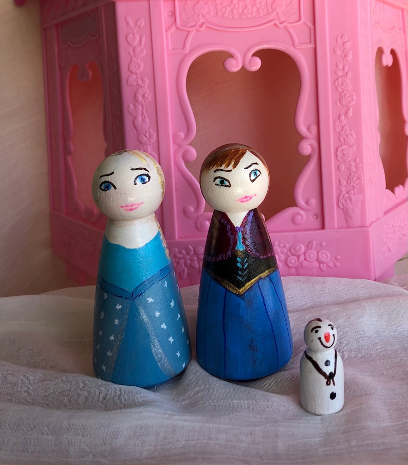 May include: Hand-painted wooden peg dolls of Elsa and Anna from the film Frozen, dressed in their signature blue and purple gowns. A small snowman peg doll is also included.