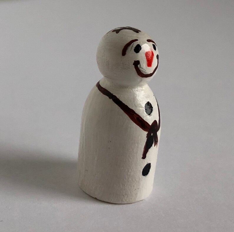 May include: A white wooden snowman figurine with a black scarf and black buttons. The snowman has a painted red nose and black eyes.