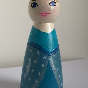 May include: A hand-painted wooden doll wearing a blue dress with a white snowflake pattern. The doll has blonde hair, blue eyes, and a pink mouth.