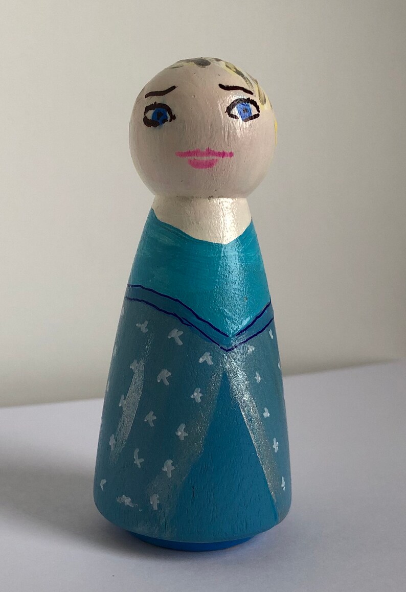 May include: A hand-painted wooden doll wearing a blue dress with a white snowflake pattern. The doll has blonde hair, blue eyes, and a pink mouth.