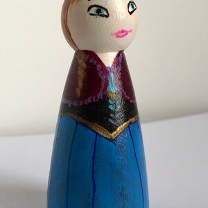 May include: Hand-painted wooden doll with brown hair, blue eyes, and a pink mouth. The doll is wearing a blue dress with a black bodice and a purple and gold trim.