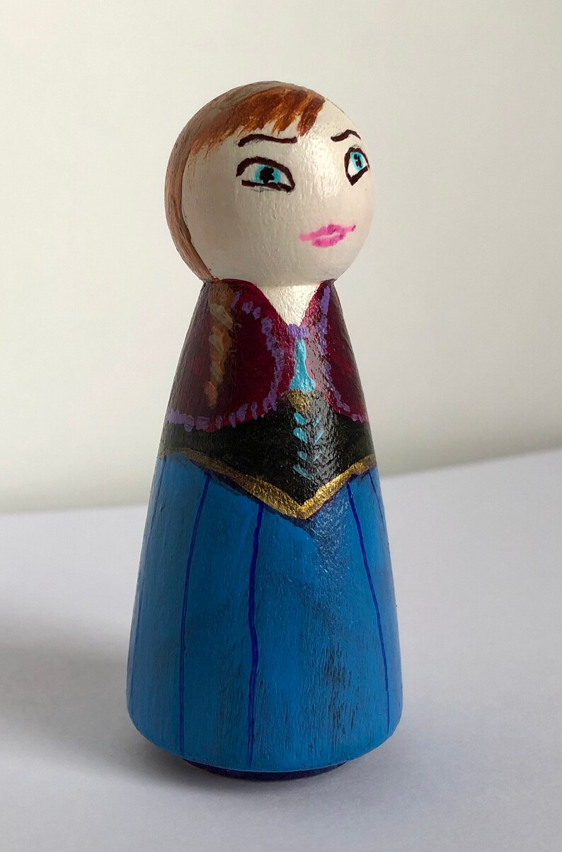 May include: Hand-painted wooden doll with brown hair, blue eyes, and a pink mouth. The doll is wearing a blue dress with a black bodice and a purple and gold trim.