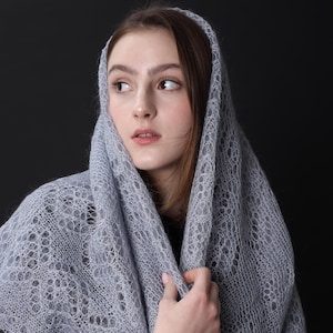Może przedstawiać: Kobieta w jasnoszarym, dzianym szalu z delikatnym wzorem koronkowym. Szal jest zarzucony na głowę i ramiona, a ona patrzy w bok.