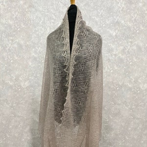 Puede incluir: Un chal delicado de color gris claro con un patrón de encaje y bordes con flecos. El chal está drapeado sobre un maniquí.