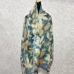 May include: A lightweight scarf with a leaf pattern in shades of blue, beige, and cream. The scarf is draped over a mannequin, showcasing the design and texture. The fabric appears to be soft and flowing.