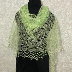 Peut inclure: Un châle vert clair avec un tissage ajouré et une bordure festonnée. Le châle est présenté sur un mannequin noir, mettant en valeur son design délicat et dentelé. La couleur est un vert pastel doux, et le matériau semble léger et aéré.