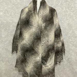 May include: A handmade, open-weave shawl in shades of gray and cream. The shawl features a scalloped edge and a gradient pattern, transitioning from dark gray to cream. The delicate knit creates a lacy effect, perfect for layering.