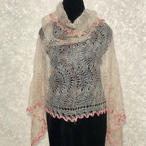 May include: A fine, open-weave shawl in beige and pale pink. The shawl has a scalloped edge in a darker pink. The garment is draped over a black mannequin, highlighting its intricate design and texture. The shawl is approximately 180 cm long.