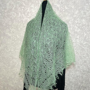 May include: A delicate, light green lace shawl with a delicate, open weave pattern. The shawl is draped over a mannequin and has a soft, flowing drape.