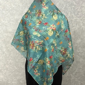 May include: A square, teal scarf with a floral pattern. The scarf features a repeating design of colorful flowers in shades of red, yellow, and white. The scarf is draped over a black garment.