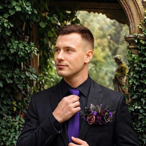 May include: A black suit jacket worn over a black button-down shirt with a textured purple necktie. A floral boutonniere featuring dark purple and mauve roses with green foliage is pinned to the left lapel of the black suit jacket.