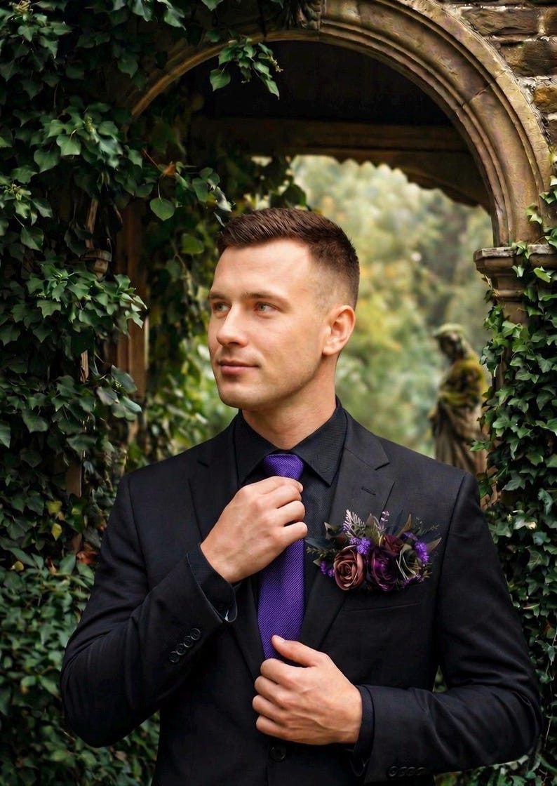 May include: A black suit jacket worn over a black button-down shirt with a textured purple necktie. A floral boutonniere featuring dark purple and mauve roses with green foliage is pinned to the left lapel of the black suit jacket.