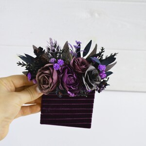 May include: A hair accessory featuring a cluster of artificial flowers in shades of deep purple, plum, and dark gray, accented with dark foliage and small purple buds, attached to a rectangular base covered in dark purple velvet ribbon strips.