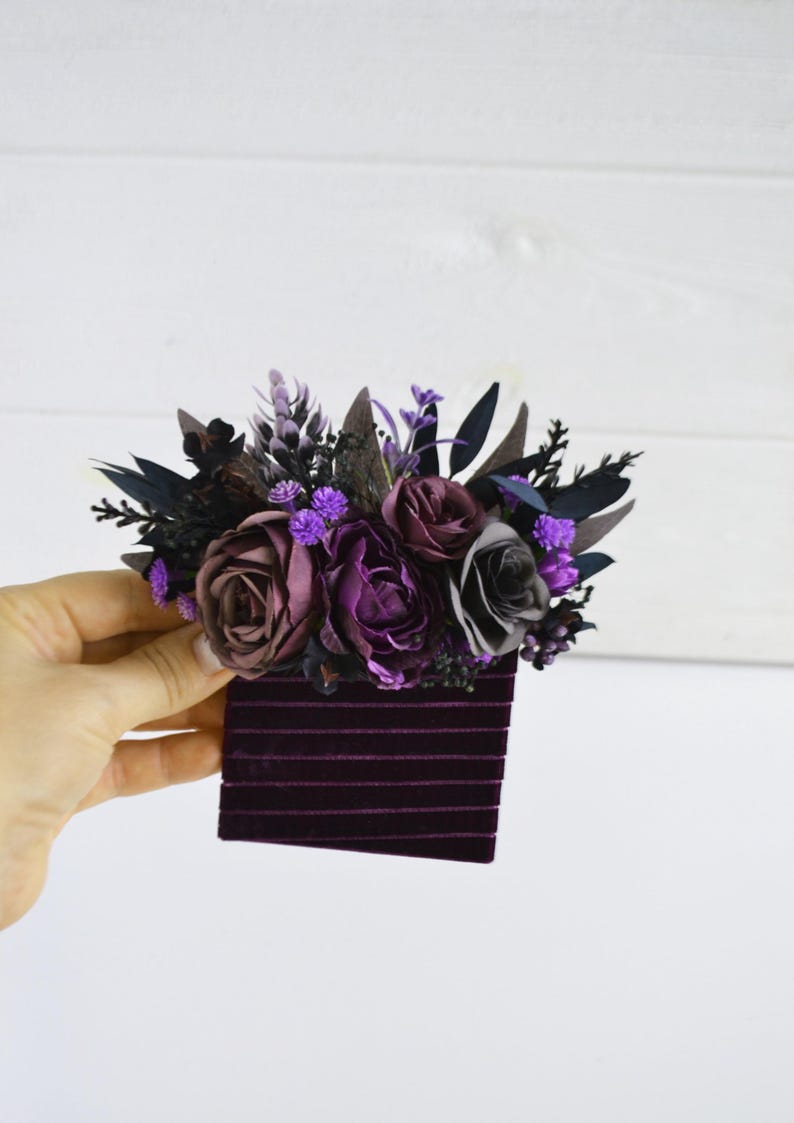 May include: A hair accessory featuring a cluster of artificial flowers in shades of deep purple, plum, and dark gray, accented with dark foliage and small purple buds, attached to a rectangular base covered in dark purple velvet ribbon strips.