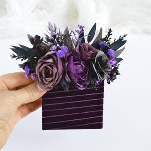 May include: A rectangular dark purple velvet hair accessory featuring a decorative arrangement of artificial flowers in shades of deep purple, lavender, and black, accented with dark green foliage and small purple sprigs.