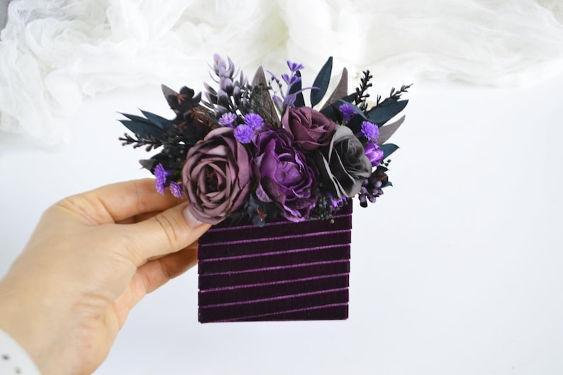 May include: A rectangular dark purple velvet hair accessory featuring a decorative arrangement of artificial flowers in shades of deep purple, lavender, and black, accented with dark green foliage and small purple sprigs.