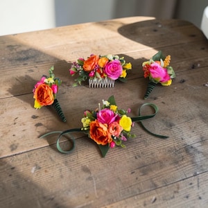May include: A collection of colorful floral accessories, including boutonnieres, a hair comb, and a wrist corsage. The flowers are in shades of orange, pink, and yellow, with green accents. The items are arranged on a rustic wooden surface.