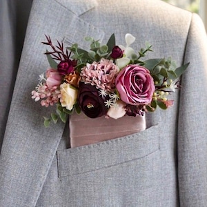 May include: A grey suit jacket with a pocket square and a floral arrangement. The flowers include burgundy, pink, and white roses, carnations, and greenery. The pocket square is a light pink colour.