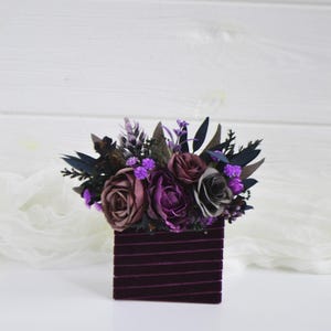 May include: A square container wrapped in dark purple velvet ribbon, topped with an arrangement of artificial flowers in shades of deep purple, mauve, and grey, accented with dark green foliage and small bright purple filler blossoms.