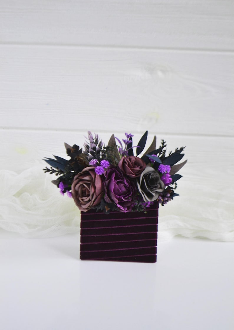 May include: A square container wrapped in dark purple velvet ribbon, topped with an arrangement of artificial flowers in shades of deep purple, mauve, and grey, accented with dark green foliage and small bright purple filler blossoms.