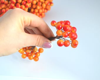 May include: A hand holding a sprig of faux berries. The berries are a mix of orange and red.