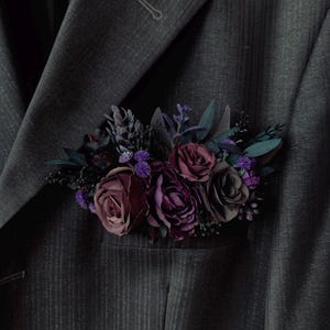 May include: A dark gray pinstriped suit jacket featuring a pocket square arrangement of artificial flowers in deep shades of burgundy, plum, and dark purple, accented with small sprigs of dark green foliage and tiny violet blossoms.