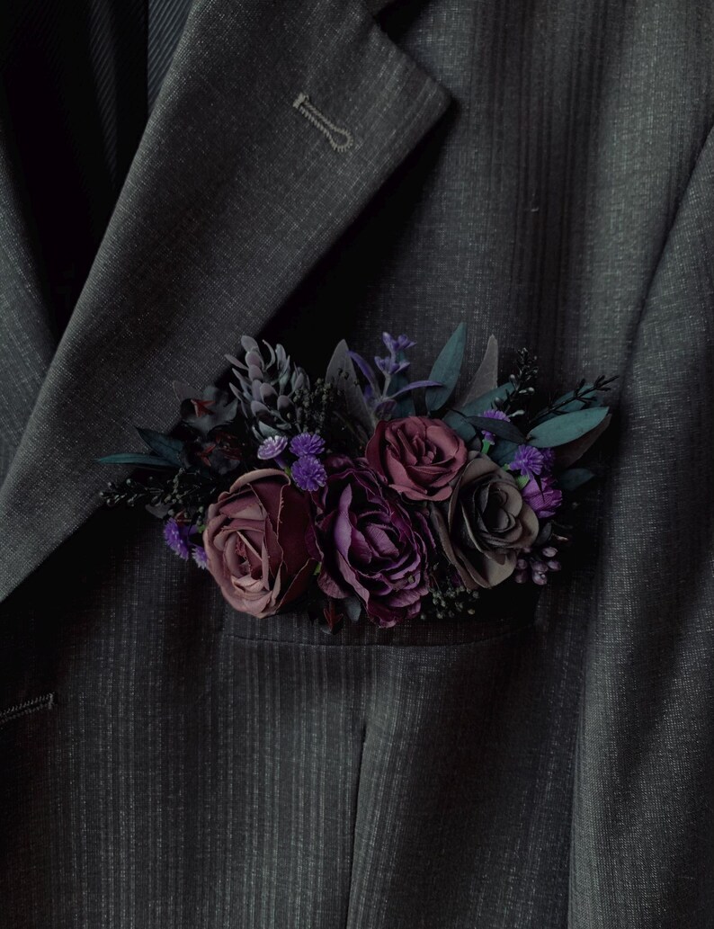 May include: A dark gray pinstriped suit jacket featuring a pocket square arrangement of artificial flowers in deep shades of burgundy, plum, and dark purple, accented with small sprigs of dark green foliage and tiny violet blossoms.