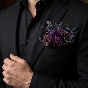 May include: A black blazer and black shirt featuring a pocket square arrangement of artificial flowers in shades of deep purple, mauve, and dark grey. The floral cluster includes roses and small filler blossoms tucked into the chest pocket.