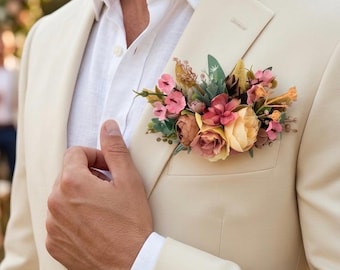 May include: A cream-colored blazer worn over a white collared shirt, featuring a floral boutonniere in the breast pocket. The arrangement includes pink, yellow, and brown artificial flowers with green foliage, pinned to the lapel area.