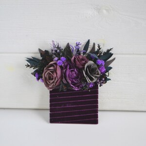 May include: A rectangular dark purple velvet base featuring horizontal ribbed texture, topped with an arrangement of artificial flowers in shades of deep purple, mauve, and grey, accented with dark green foliage and small bright purple filler blooms.
