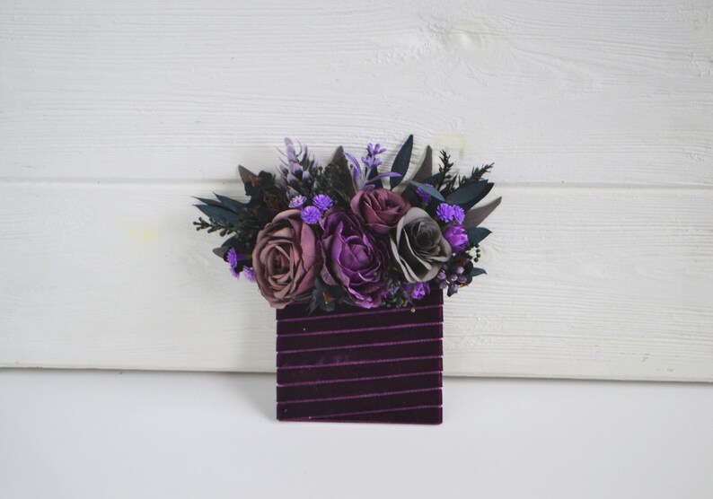 May include: A rectangular dark purple velvet base featuring horizontal ribbed texture, topped with an arrangement of artificial flowers in shades of deep purple, mauve, and grey, accented with dark green foliage and small bright purple filler blooms.