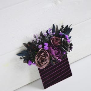 May include: A decorative accessory featuring a cluster of artificial flowers in shades of deep purple, mauve, and black, accented with dark foliage. The floral arrangement is attached to a rectangular base made of dark purple velvet with thin stripes.