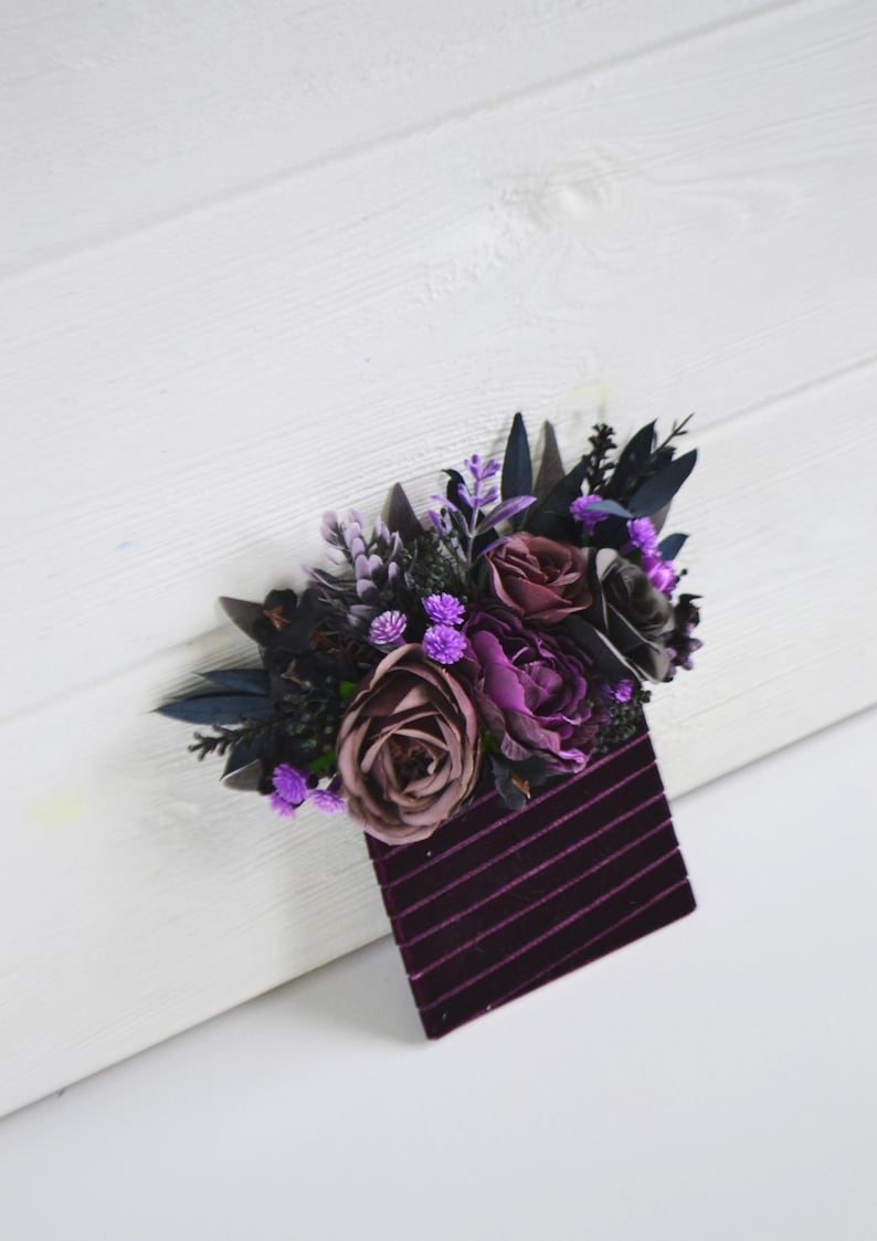 May include: A decorative accessory featuring a cluster of artificial flowers in shades of deep purple, mauve, and black, accented with dark foliage. The floral arrangement is attached to a rectangular base made of dark purple velvet with thin stripes.