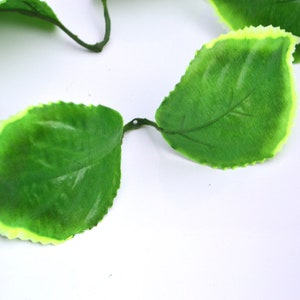 May include: A garland of artificial green leaves with a light yellow border. The leaves are arranged in a chain and have a realistic texture.