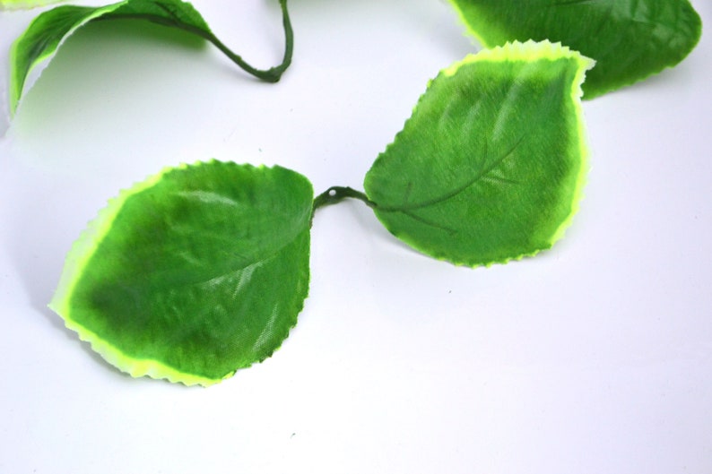 May include: A garland of artificial green leaves with a light yellow border. The leaves are arranged in a chain and have a realistic texture.