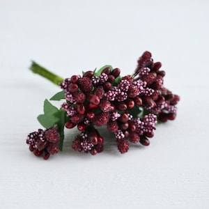 May include: A bunch of burgundy artificial berry sprigs with small pink accents. The berries are clustered together and have a slightly glittery finish.