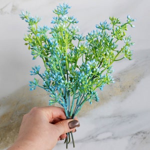 Pode incluir: Um ramo de flores artificiais azuis e verdes com pequenas flores delicadas. As flores estão dispostas em um ramo e têm um aspecto natural e realista.