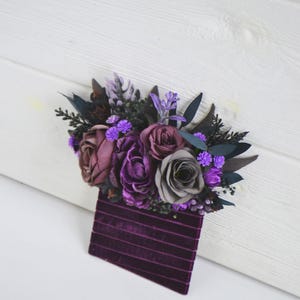 May include: A boutonniere featuring a cluster of artificial flowers in shades of deep purple, plum, and grey, accented with dark foliage and small bright purple blossoms, attached to a rectangular base of dark purple velvet ribbon with horizontal lines.