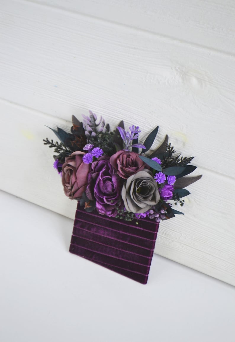 May include: A boutonniere featuring a cluster of artificial flowers in shades of deep purple, plum, and grey, accented with dark foliage and small bright purple blossoms, attached to a rectangular base of dark purple velvet ribbon with horizontal lines.
