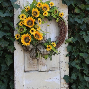 May include: A rustic grapevine wreath adorned with vibrant yellow sunflowers, green leaves, and a black and white checkered bow. The wreath hangs on a distressed, off-white door, with ivy in the background.