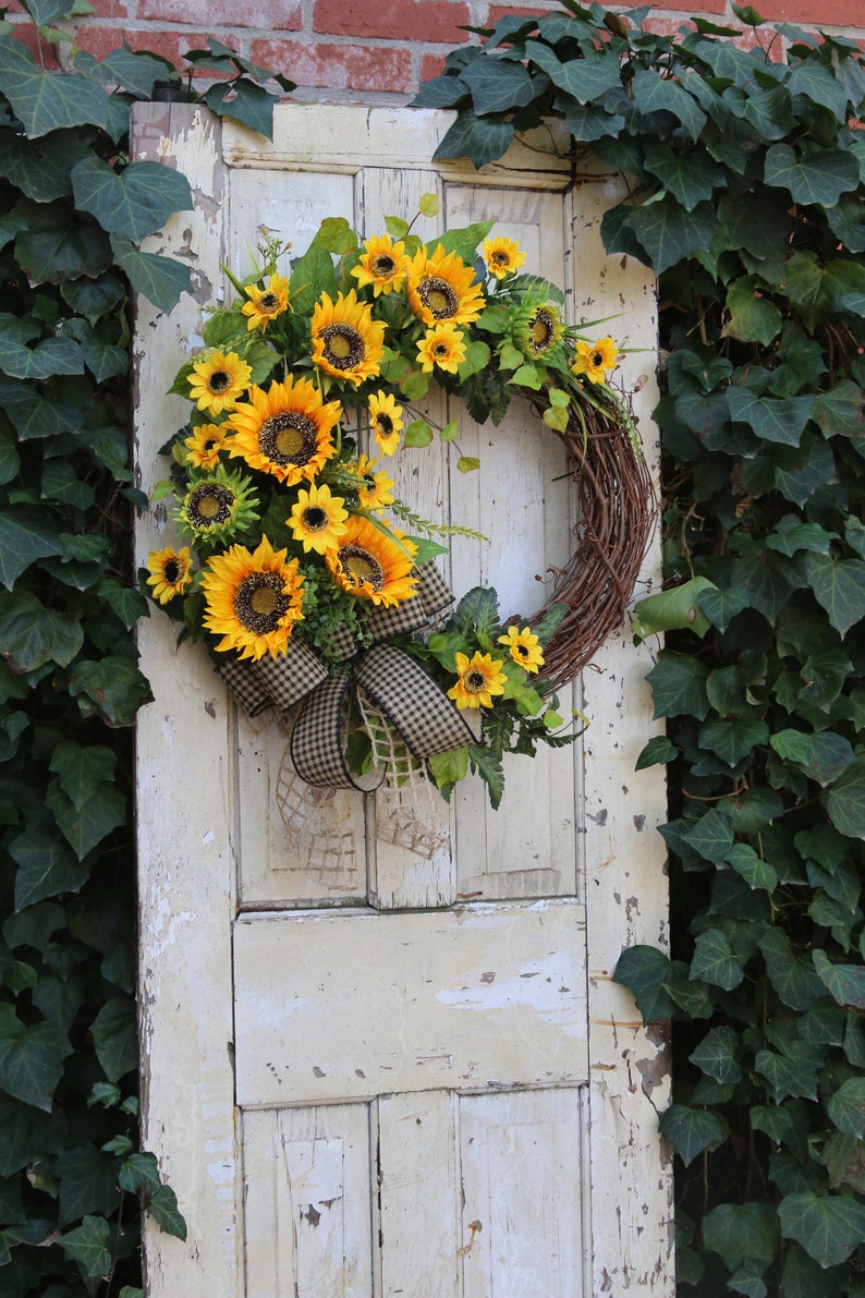 May include: A rustic grapevine wreath adorned with vibrant yellow sunflowers, green leaves, and a black and white checkered bow. The wreath hangs on a distressed, off-white door, with ivy in the background.