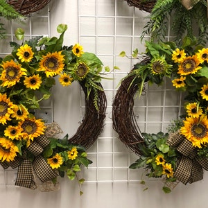 May include: Two decorative wreaths with a rustic, woven base. Each wreath is adorned with vibrant yellow sunflowers, green leaves, and a checkered bow. The wreaths are displayed against a white grid background.