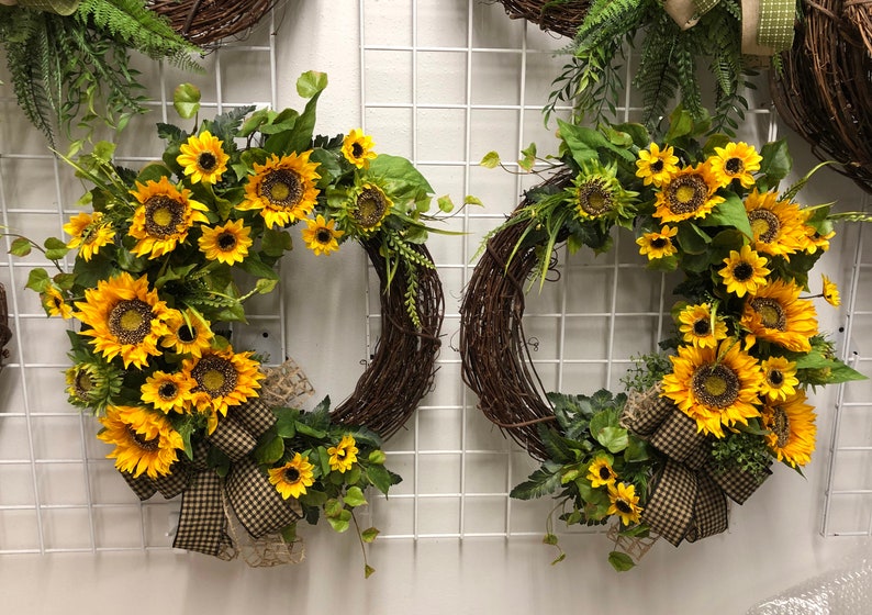 May include: Two decorative wreaths with a rustic, woven base. Each wreath is adorned with vibrant yellow sunflowers, green leaves, and a checkered bow. The wreaths are displayed against a white grid background.