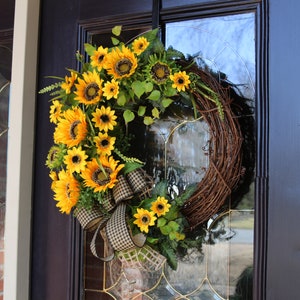 May include: A decorative wreath featuring bright yellow sunflowers and green leaves arranged on a natural grapevine base. A large bow with a black and white checkered pattern adds a decorative touch. The wreath hangs on a dark door.