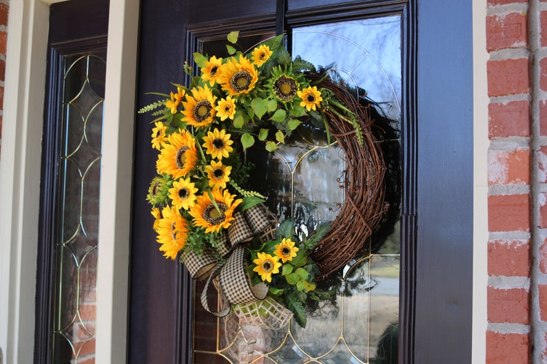 May include: A decorative wreath featuring bright yellow sunflowers and green leaves arranged on a natural grapevine base. A large bow with a black and white checkered pattern adds a decorative touch. The wreath hangs on a dark door.