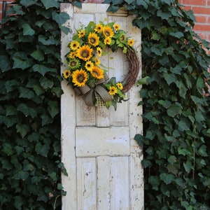 May include: A rustic, weathered, off-white door is adorned with a vibrant sunflower wreath. The wreath features bright yellow sunflowers, green foliage, and a brown and white checkered bow. The door is set against a backdrop of lush green ivy and a red brick wall.