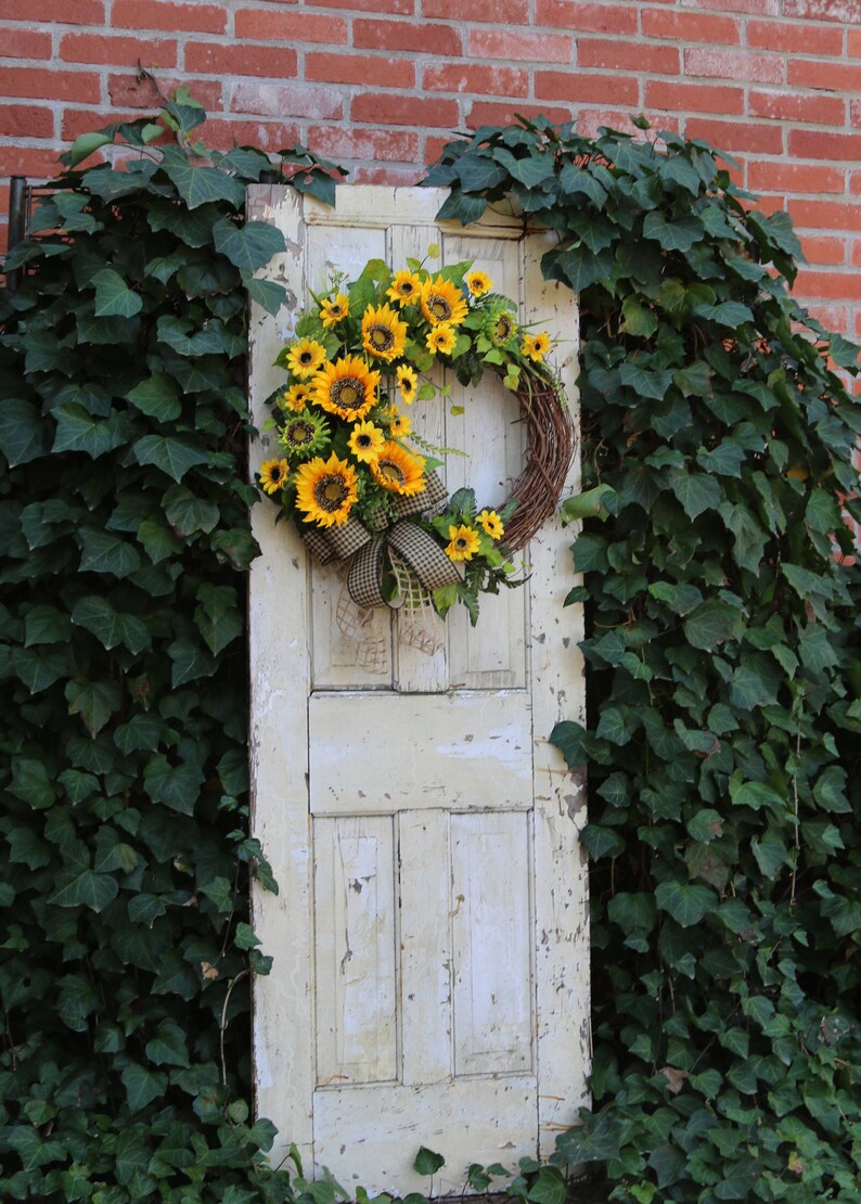 May include: A rustic, weathered, off-white door is adorned with a vibrant sunflower wreath. The wreath features bright yellow sunflowers, green foliage, and a brown and white checkered bow. The door is set against a backdrop of lush green ivy and a red brick wall.