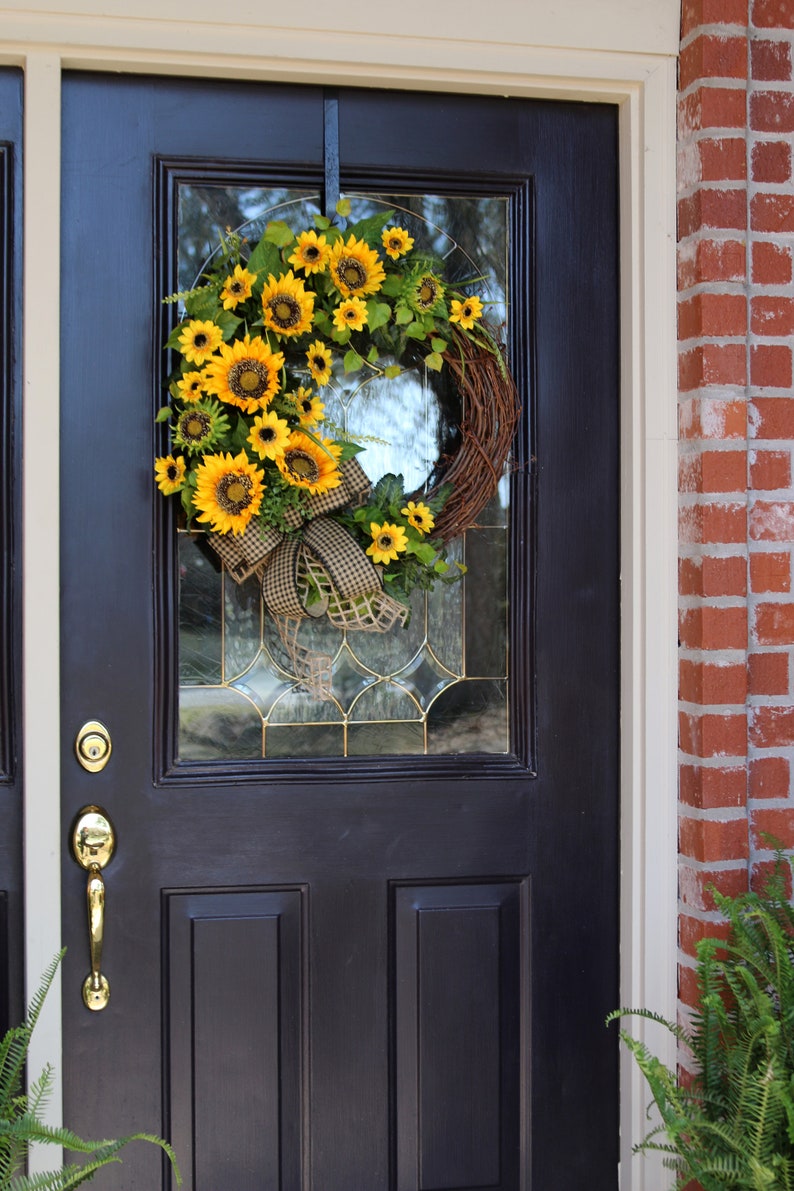 May include: A decorative sunflower wreath hangs on a black front door. The wreath is made of woven twigs and features bright yellow sunflowers, green leaves, and a brown and white checkered bow. The door has a gold handle and a glass panel.
